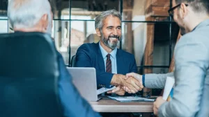 A smiling financial advisor shaking hands with a small business client across a desk with financial documents and a laptop, representing an outsourced CFO partnership and strategic business advisory.